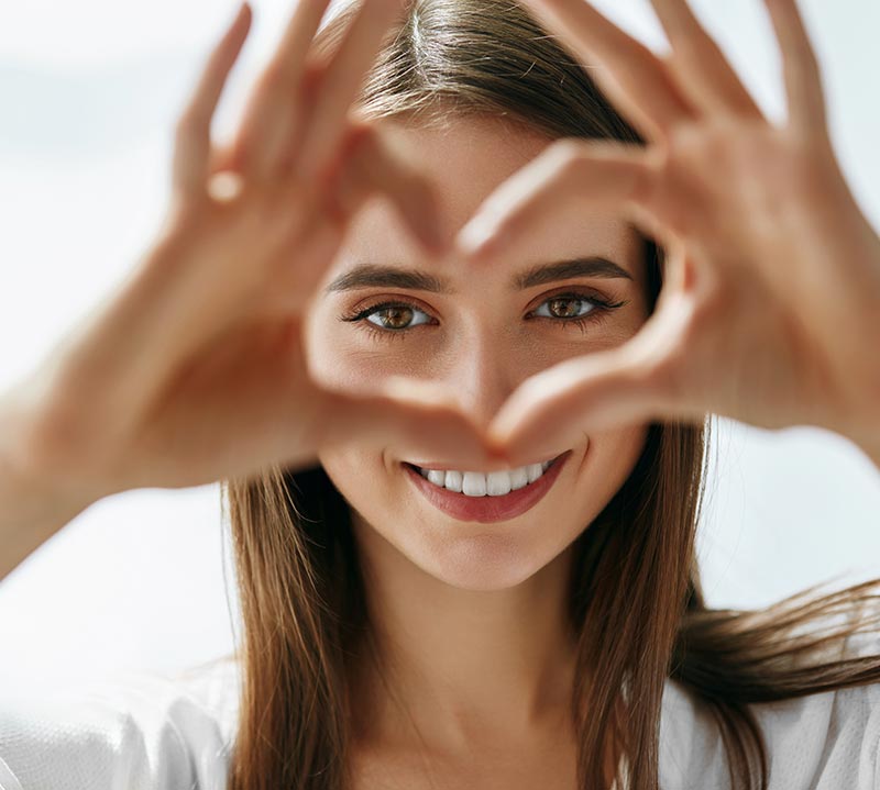 A woman looking at the camera through her hands, forming a heart with the thumbs and index fingers